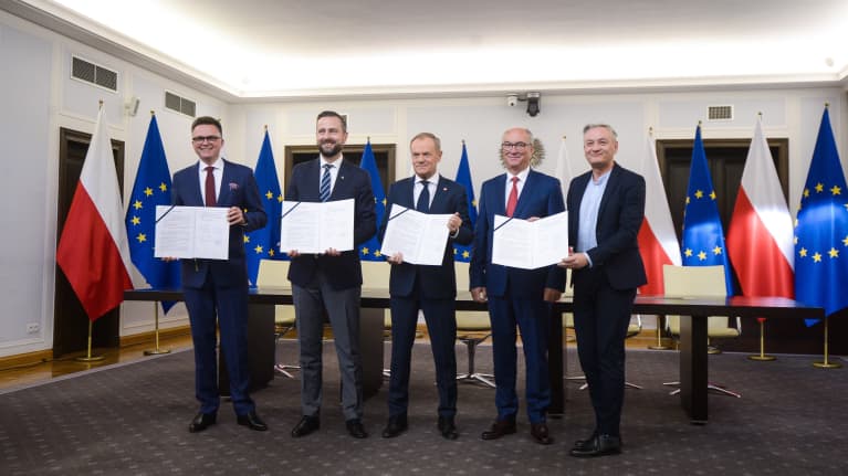 Viisi pukumiestä poseeraa kameralle Puolan ja EU:n lippurivistön edessä. He levittelevät käsissään puoluohjelmapapereita.