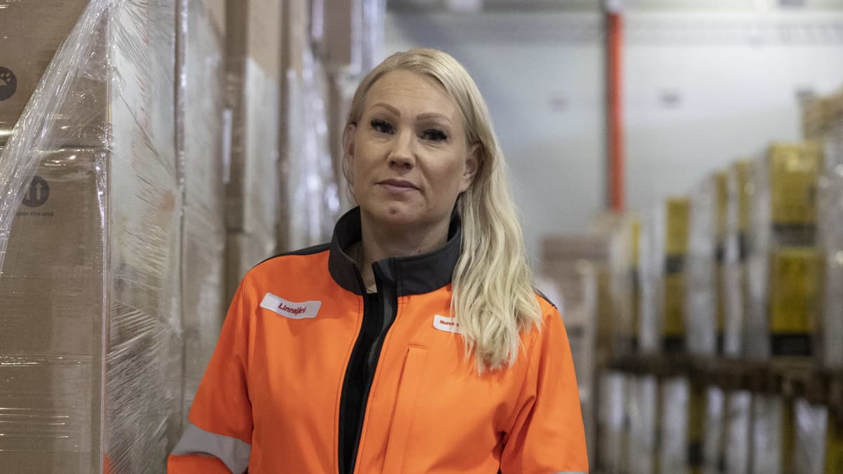 Nurminen Logistics Oyj:n myyntijohtaja Marjut Linnajärvi seisoo oranssi huomiotakki päällä satamavarastolla ja katsoo suoraan kameraan.