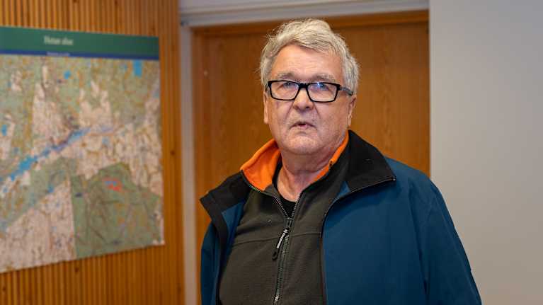 Henkilö seisoo sisätilassa kartan edessä. Hänellä on yllään tummansininen takki ja tumma paita, jossa on oranssia kauluksessa.