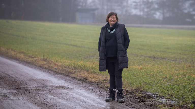 Maria Wasström står på en grusväg vid sidan av ett fält en dimmig dag. Hon bär en svart klädsel och ett halsband.