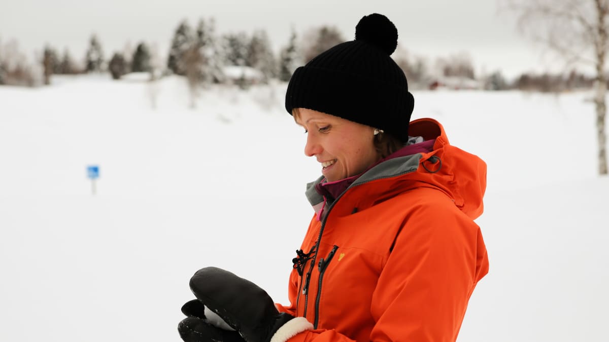 Kaisa Sali lumisessa maisemassa. Kaisalla on punainen kuoritakki ja musta pipo. Käsissään hän muotoilee lumipalloa. Taustalla on jäätynyt joen uoma, vastarannalla on metsää ja mökkejä. Keskellä jokea on sininen kyltti, josta ei saa kuvassa selvää.