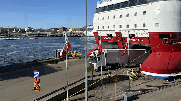 Lorry driving out of the stern of a red and white ro-ro ferry, with a harbour, part of another ferry and the cityscape in the background.