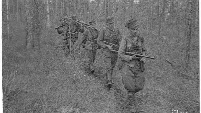Mustavalkoisessa kuvassa neljä maailmansodan aikaisessa suomalaisessa asepuvussa olevaa miestä marssii suometsässä ja jokaisella on ase mukanaan. 