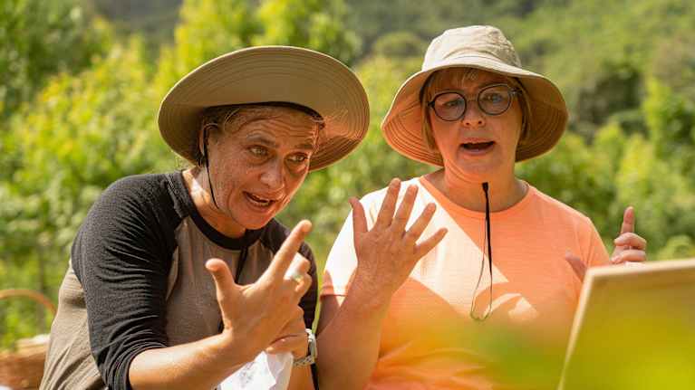 Aino-Kaisa Pekonen ja Silvia Modig vaaleat Brimmer-hatut päässään lukevat jotain edessä olevalta taululta ulkona. Taustalla vehreitä puita.