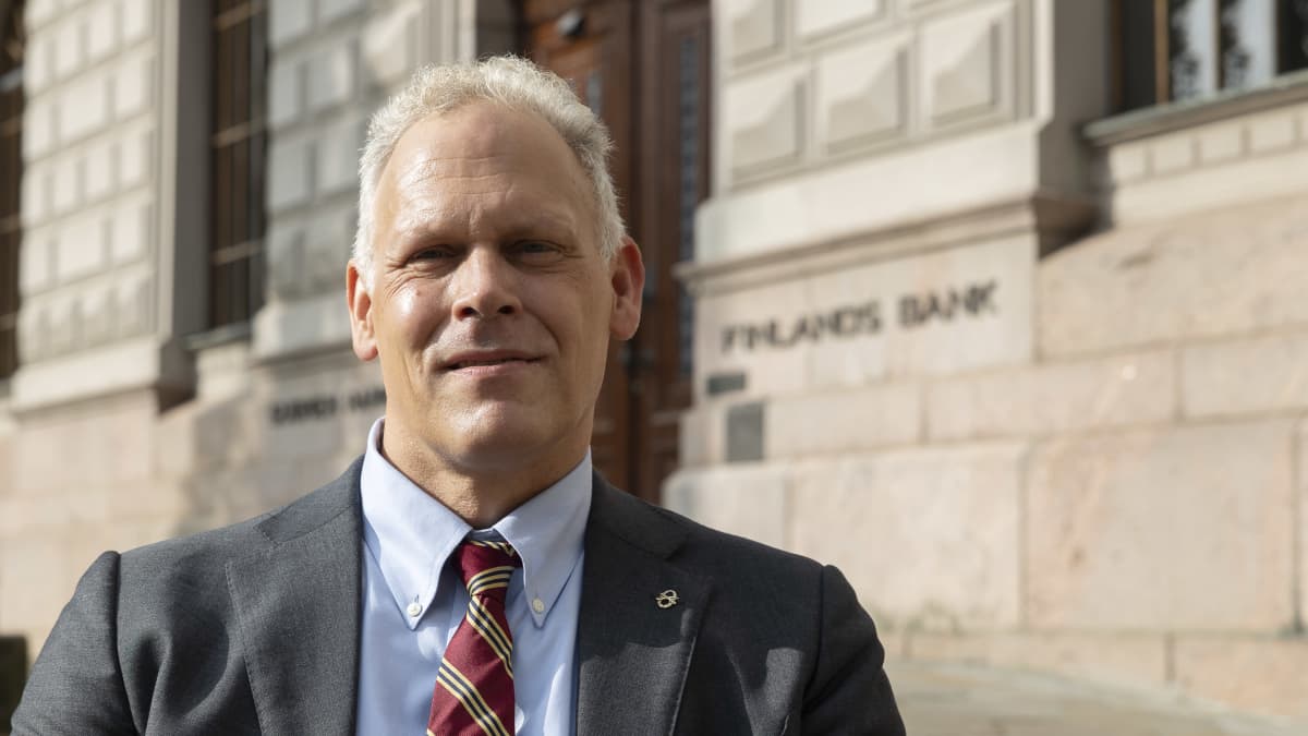 Pukumies seisoo kivitalon edustalla ja katsoo kameraan.