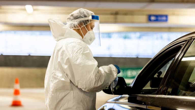 Person in white coveralls and face mask and clear shield administering a Covid test to someone sitting in a black car in a large parking garage.