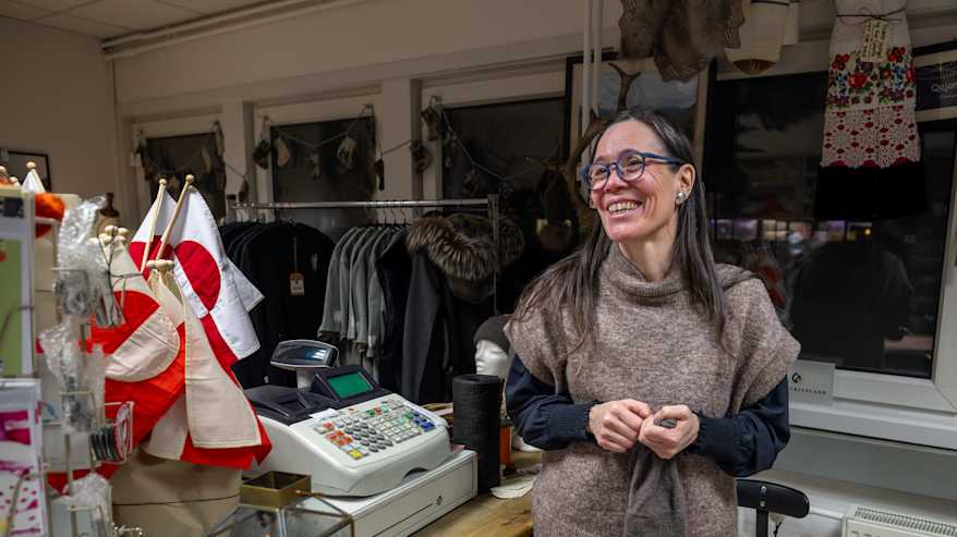 Tumma, pitkähiuksinen nainen, Vivi Schultz, puodissaan kassan takana. Neoulos kädessään. Hymyilee.