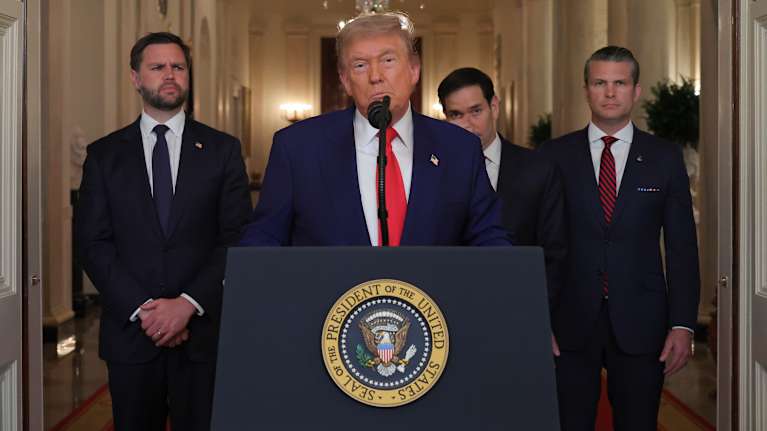 Presidentti Donald Trump sinisessä puvussa ja punaisessa solmiossa puhujapöntössä. Takana kolme miestä puvuissa.