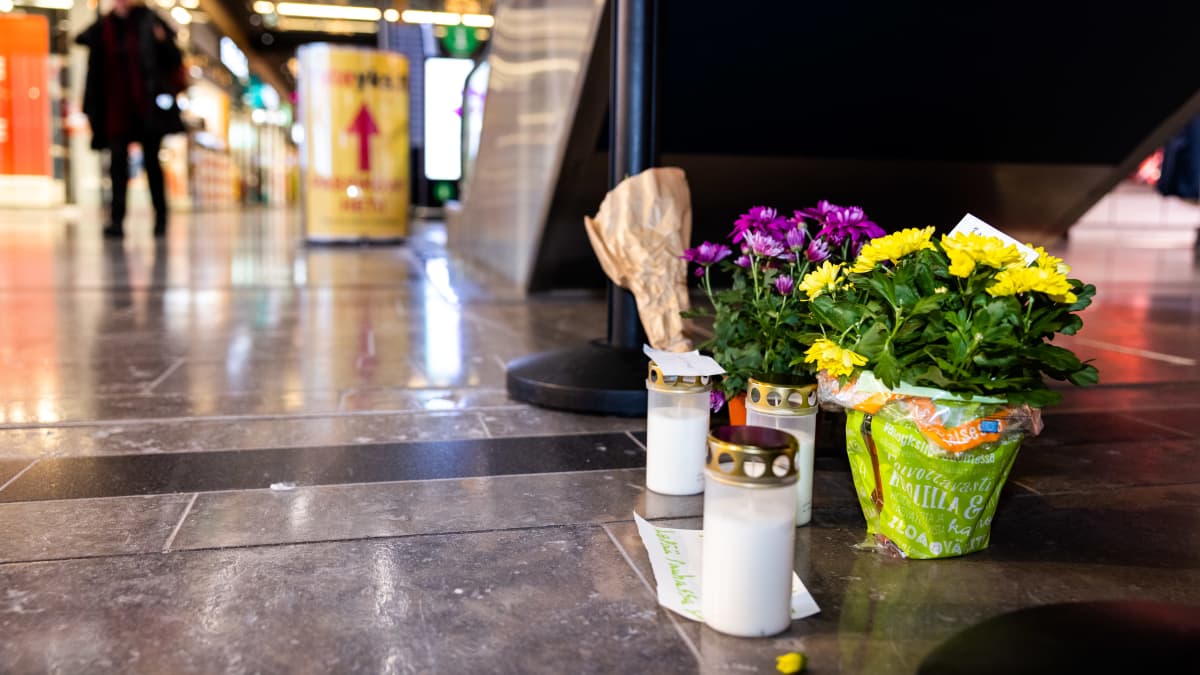 Interior shot of a shopping mall with a few grave candles and flowers on the floor to the right and a dim human figure in the distance on the left.