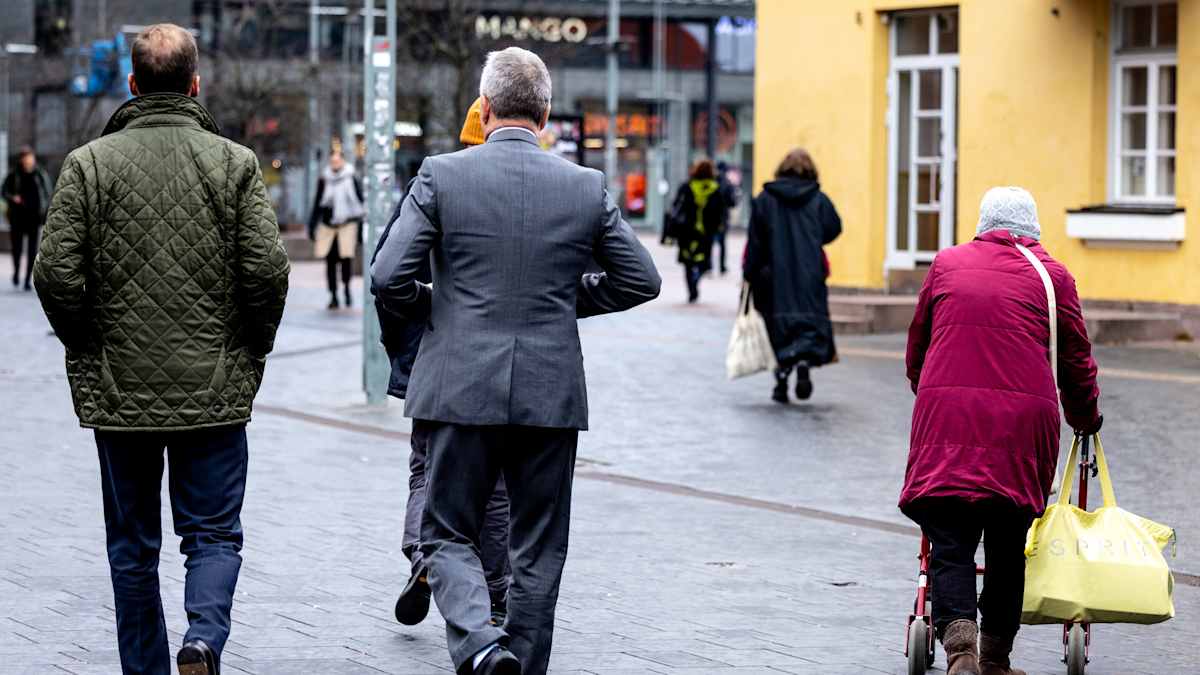 Vanhus rollaattorin kanssa ja kaksi pukuun pukeutunutta henkilö kadulla.