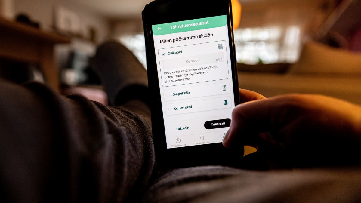 Puhelin applikaatiolla henkilö tilaamassa kotiinkuljetuspalvelua istuen tohvelit jalassa asunnossa.