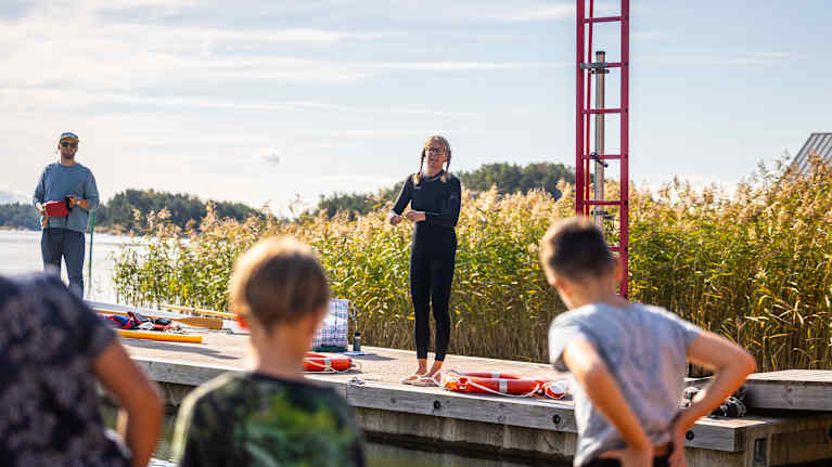 Lapset kuuntelevat ohjeita laiturilla aurinkoisena päivänä, taustalla kauniit kaislikot ja sininen taivas.