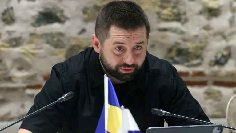 Davyd Arahamija puhuu pöydän ääressä edessään näyttö ja mikrofoneja. Hänellä on parta ja musta lyhythihainen pusakka. Etualalla näkyvä Ukrainan ja Venäjän liput.