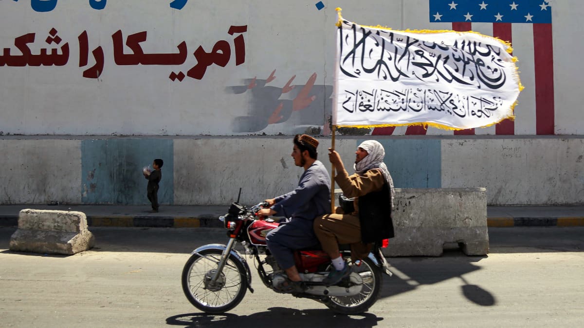 Kaksi miestä heiluttaa lippua ajaessaan moottoripyörässä entisen Yhdysvaltojen suurlähetystön edessä Kabulissa.