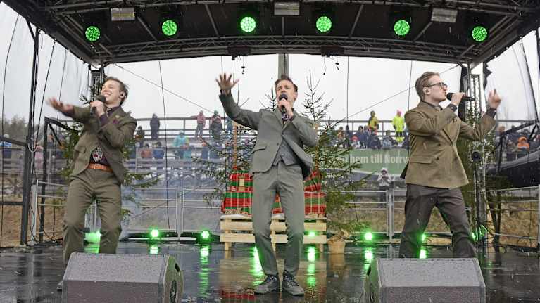 Three young men in light-brown suits waving their arms on a small damp outdoor stage with green lights, fir trees and a grey sky behind them.