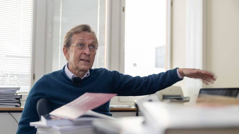 Vanhempi herrasmies istuu työpöydän takana ja viittilöi kädellään.