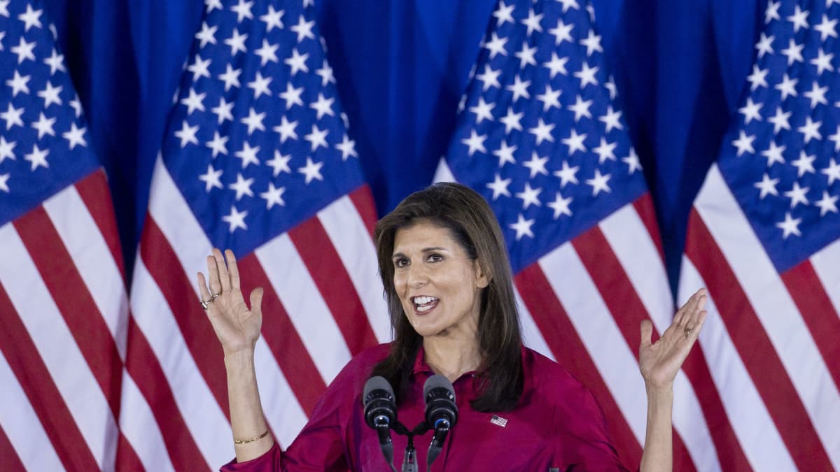 Nikki Haley speaks into a microphone with arms outstretched against a backdrop of American flags.