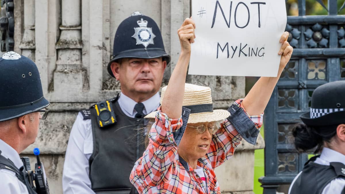 En kvinna i rutig skjorta och halmhatt håller upp en skylt där det står "Not my king", Inte min kung. Omkring henne står flera poliser, ett par i traditionella London-polishjälmar, men ingen av dem rör vid kvinnan.