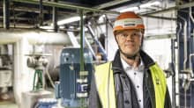 Oranssiin kypärään, nahkatakkiin ja huomioliiviin pukeutunut Olli Sirkka katsoo kameraan. Taustalla Hanasaaren voimalaitoksen laitteita ja putkia.