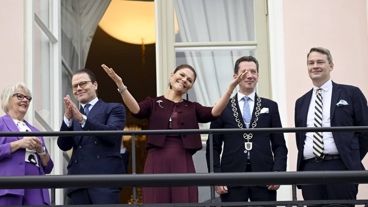 Kronprinsessan Victoria och prins Daniel på Rådhusets balkong i Lovisa tillsammans tre andra personer, en kvinna och två män. Daniel applåderar och Victoria sträcker ut armarna i uppskattning av alla de människor som samlats på torget nedanför stadshuset.