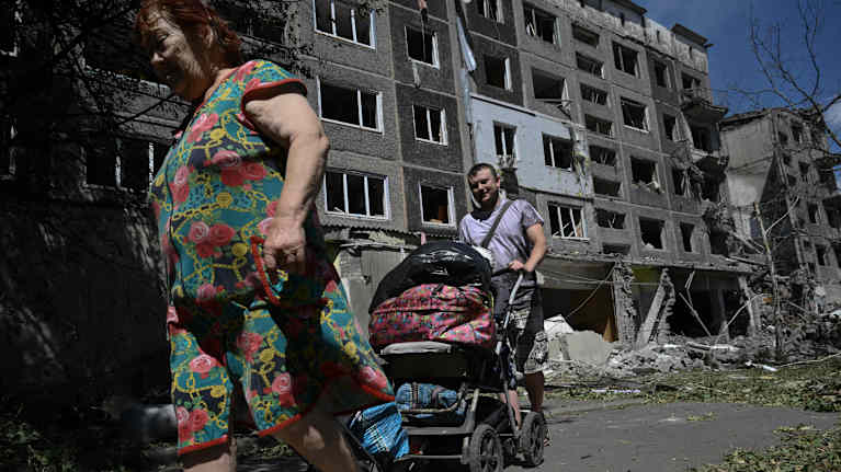 A man and a woman walk in front of a bombed house with prams. There is a lot of stuff in the carriages.