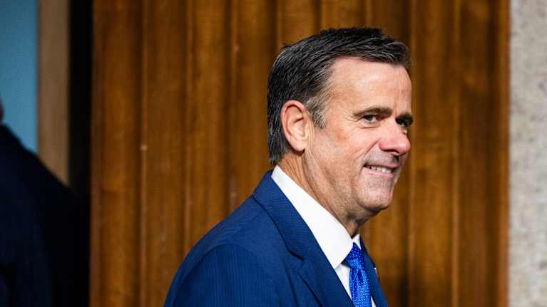 John Ratcliffe pukeutuneena siniseen pukuun ja siniseen solmioon seisoo puisessa ympäristössä.