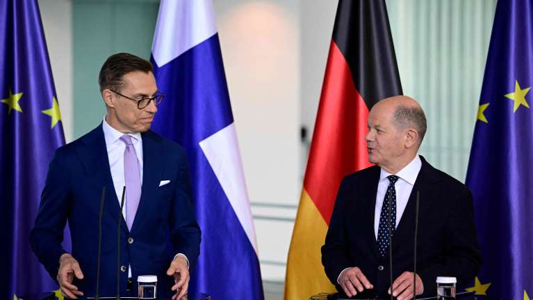  Alexander Stubb ja Olaf Scholz katsovat toisiaan, vierekkäin puhujakorokkeilla puvut päällä. Taustalla EU:n, Suomen ja Saksan liput.