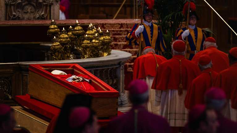 Paavi Franciscus sisältä punaiseksi verhoillussa arkussa. Takana palaa kynttilöitä. Vierellä kardinaaleja punaisissa asuissa.