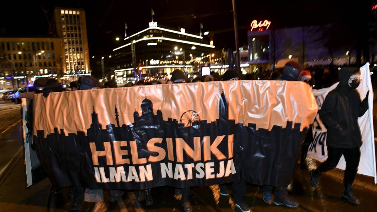 Demonstrationståget Helsingfors utan nazister marscherar i ett mörkt Helsingfors på självständighetsdagen 2021. I bakgrunden syns bland annat Glaspalatset och Posthuset.