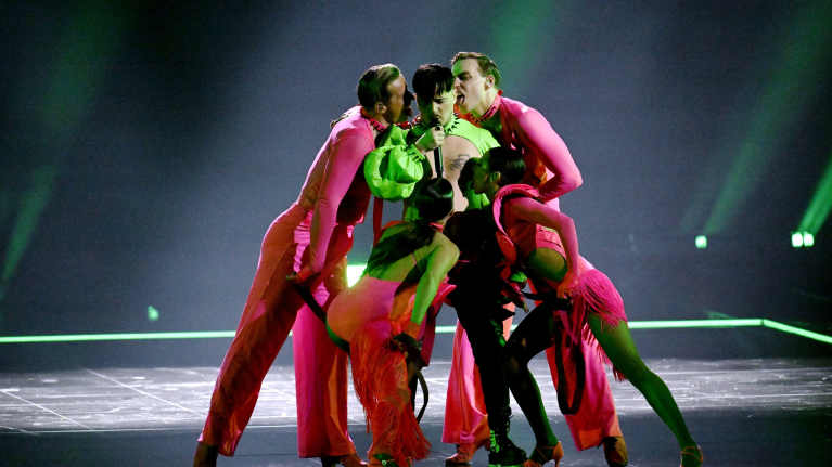 Käärijä vihreässä polerossa lavalla neljän pinkkiin pukeutuneen tanssijan ympäröimänä, jotka ovat nuolevinaan Käärijää.
