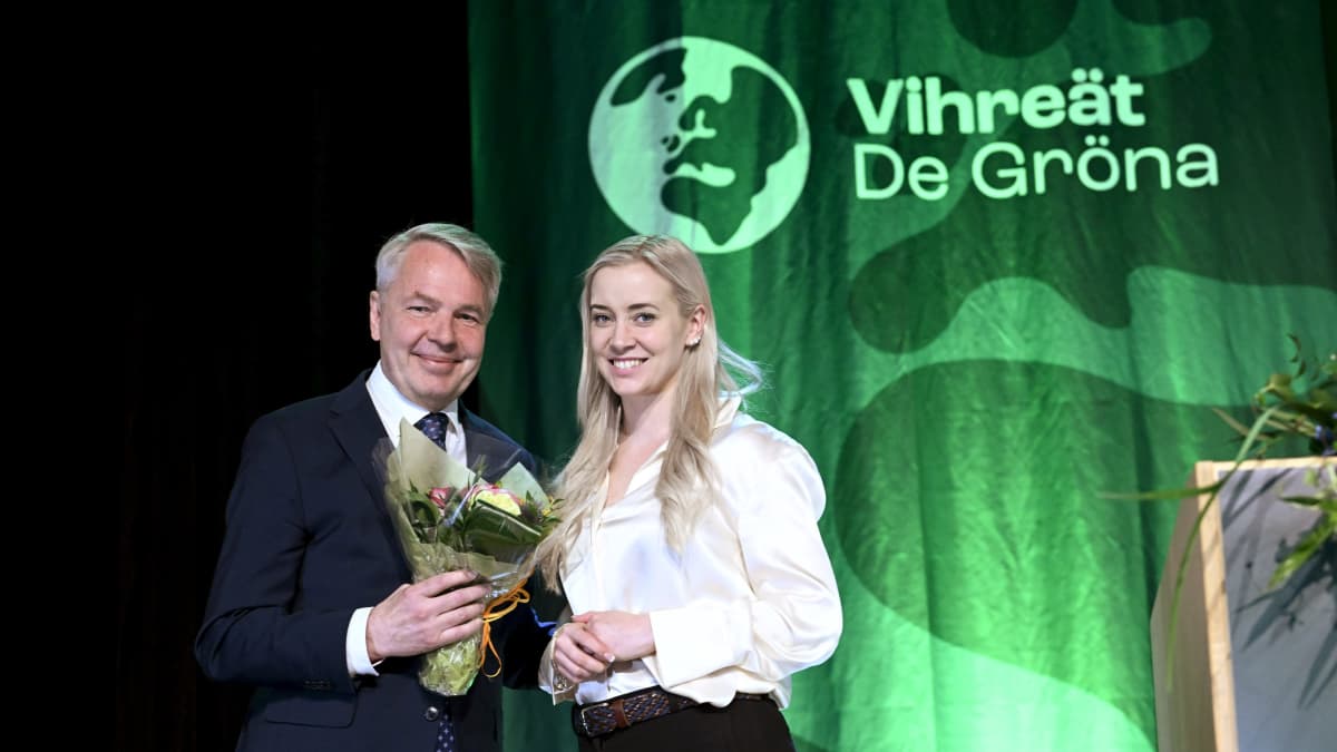 Pekka Haavisto och Sofia Virta leende tillsammans framför en grön banderoll med partiet De Grönas namn och logo.
