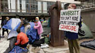 Young people setting up tents while one stands on the right holding a sign saying "UNIVERSITY YOU CAN'T HIDE YOU'RE SUPPORTING GENOCIDE"
