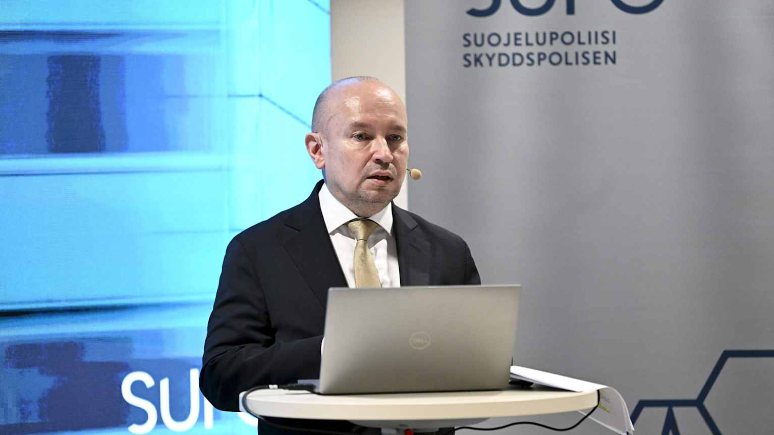 Juha Martelius puhuu korkean pöydän ääressä tietokone edessään. Takana banderolli, jossa lukee SUPO.