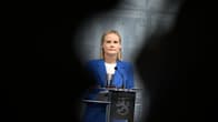 Riikka Purra in a blue jacket stands behind a podium with the emblem of the Finnish coat of arms. 