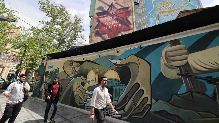 Kolme miestä kävelee Israelin vastaisen muraalin ohi Iranin pääkaupungissa Teheranissa. Muraalissa pitkäkyntiset kädet jotka puristavat moskeijaa.