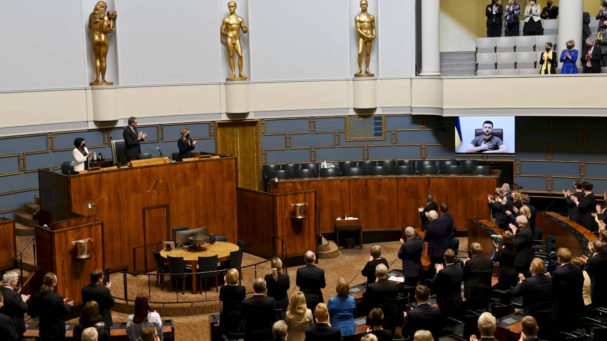 Bild från riksdagen då president Zelenskyj på distans håller tal, via en skärm. Riksdagen står och apploredar.
