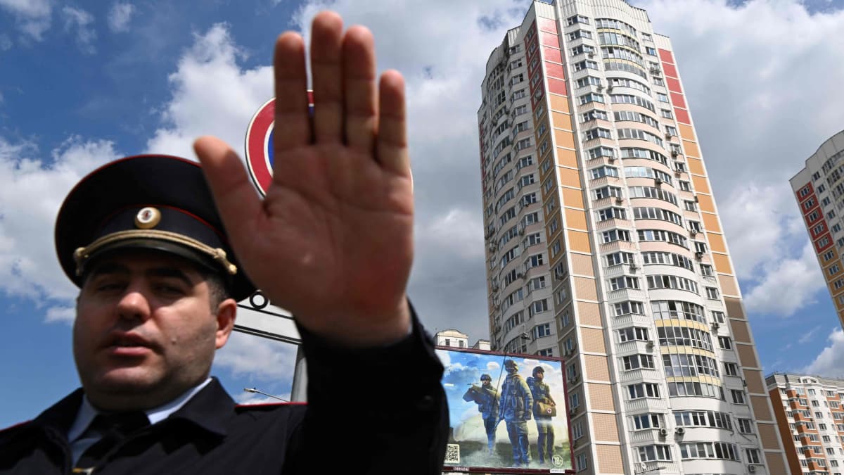 Poliisi nostaa kättään kameran eteen suuren tornitalon edessä.