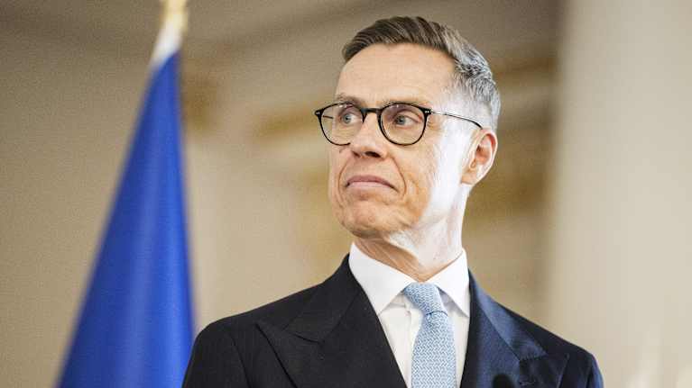 Alexander Stubb is dressed in a suit and tie. He is standing in front of the EU flag.