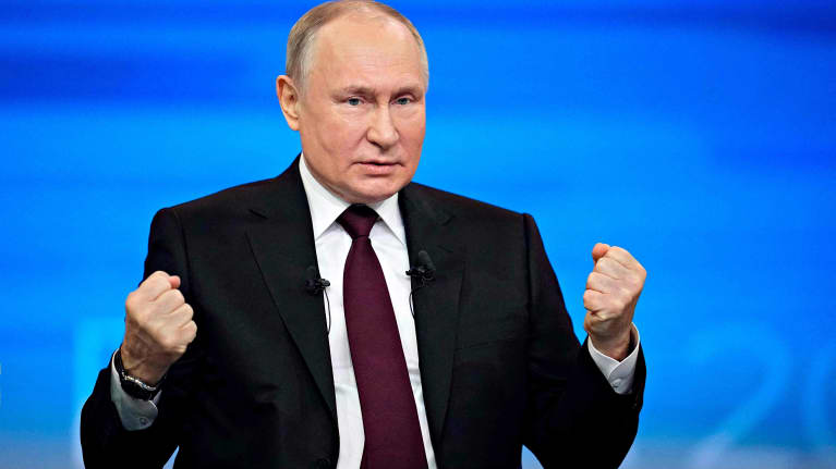 A balding man in a black jacket, white shirt and maroon tie holds up his fists while sitting behind a desk with papers.