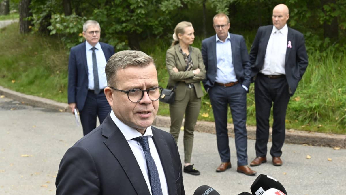 Petteri Orpo speaks in front of microphones outdoors while three men in dark suits and a woman in a light-brown suit stand in the background, with grass and bushes behind them.