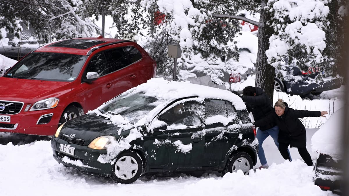 Kaksi henkilöä työntää autoa lumisessa maisemassa.