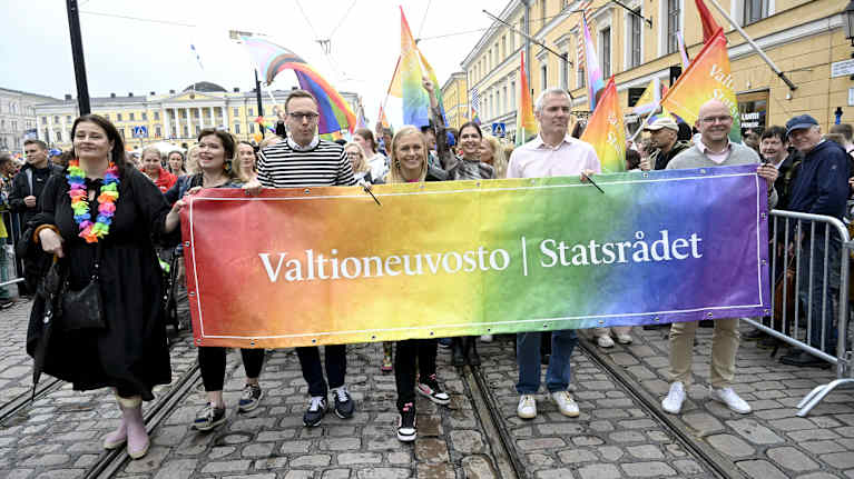 Osallistujat kantavat sateenkaaren väreissä olevaa "Valtioneuvosto | Statsrådet" -banneria Pride-kulkueessa.