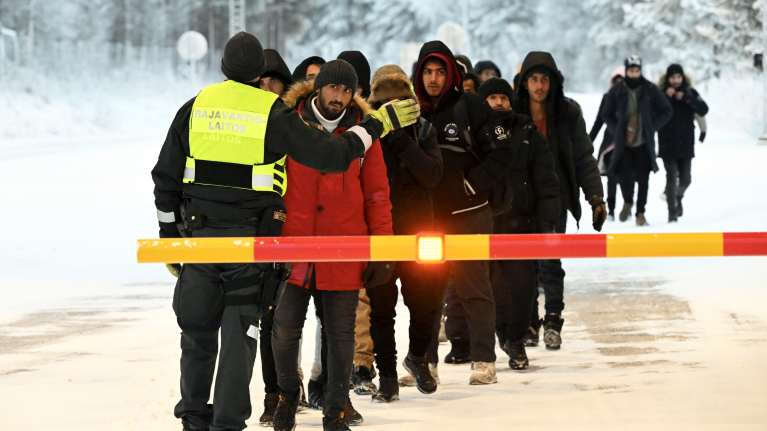 Ihmiset seisovat jonossa rajavartian edessä lumisessa maisemassa.