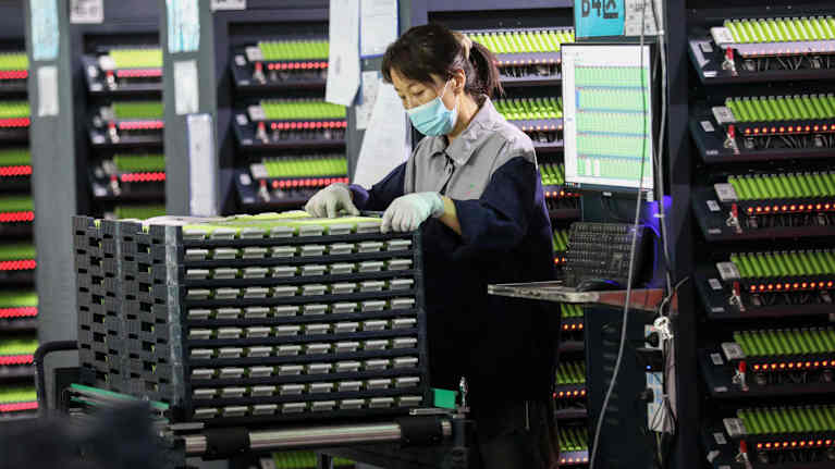 Female factory worker in uniform and face mask standing next to shelves filled with batteries.