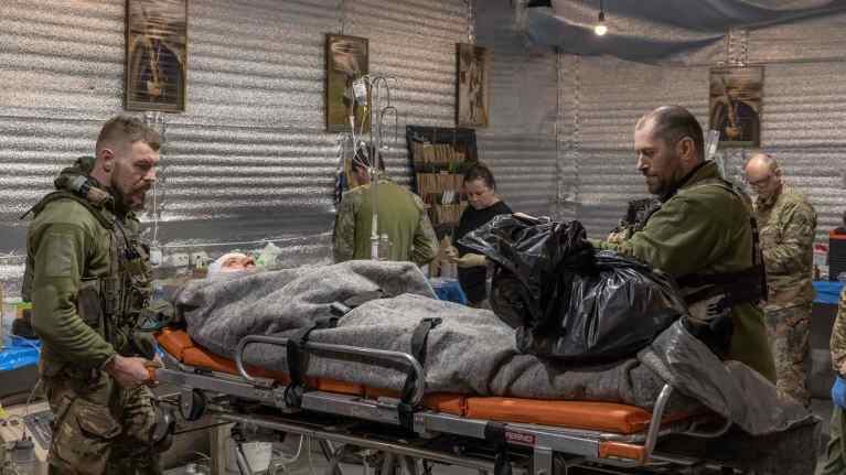 Sotilasvarusteisiin pukeutuneet lääkärit seisovat sairaalapaareilla makaavan huopaan käärityn potilaan vieressä.,