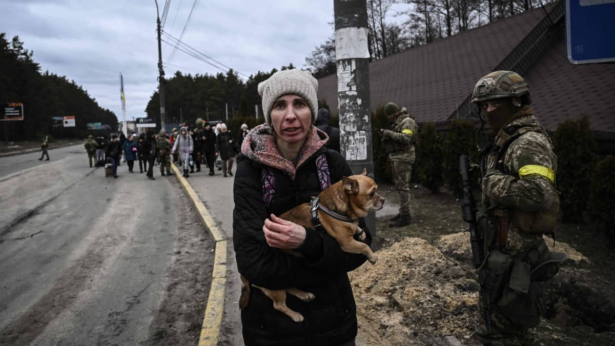 Nainen pitelee koiraa sylissä sotilas vierellään ja joukko muita evakuoitavia ihmisiä taustalla Irpinissä.