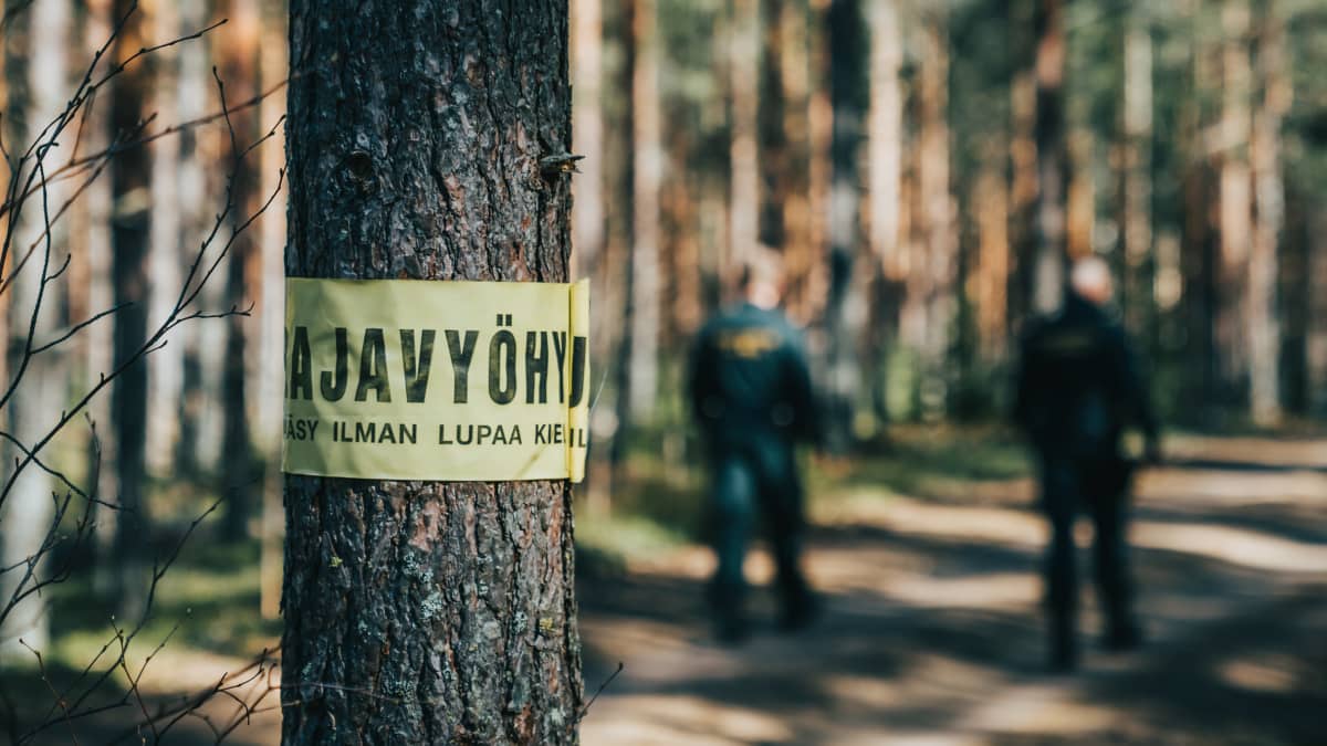A yellow ribbon is attached to a tree, which reads "no trespassing without permission". In the background, two people are walking along a forest path.