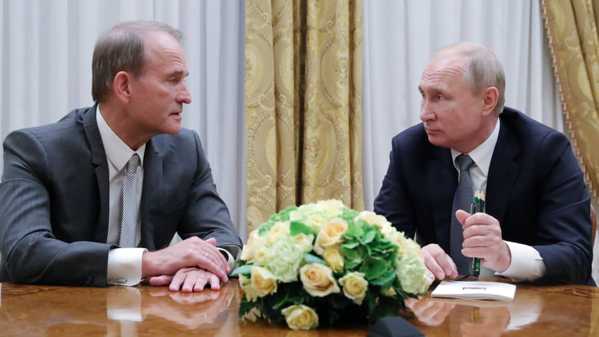 Medvedtshuk ja Putin istuvat pöydän ääressä. Medvedtshuk puhuu ja Putin kuuntelee. Medvedtshuikilla on tummanharmaa puku, Putnilla tumma. Päydällä heidän keskellään on kukka-asetelma.