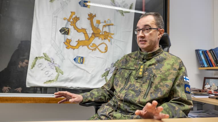 Maastopukuun pukeutunuta kadettikoulun johtaja, eversti Jukka Nurmi istuu työpöydän ääressä. Taustalla lasivitriinissä kadettikoulun suuri valkoinen lippu, jonka keskellä keltainen leijona-vaakuna.