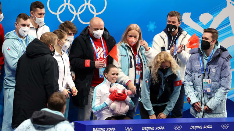 Kamila Valijeva istuu joukkuetovereidensa ympäröimänä pehmolelu sylissään.